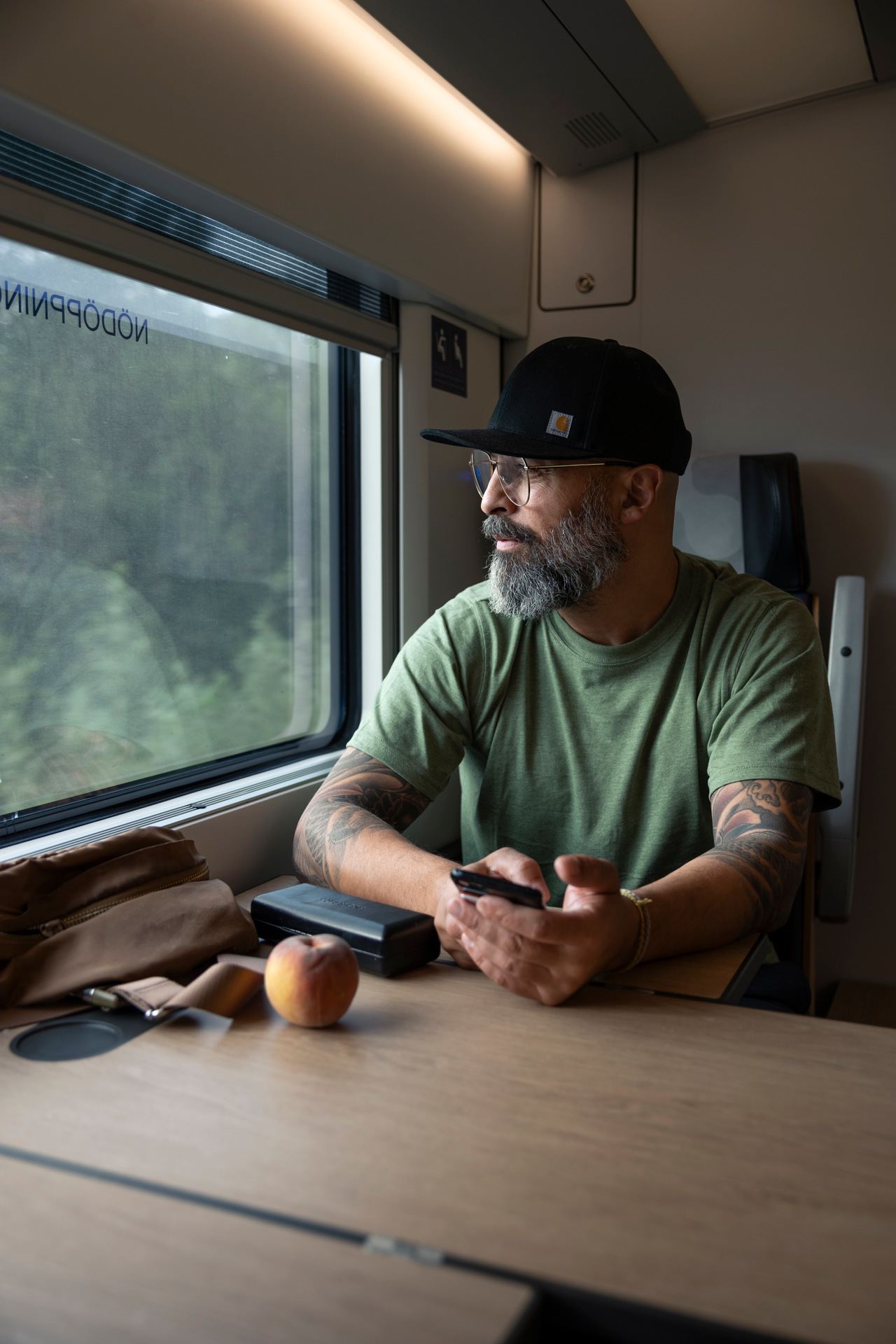 En man med glasögon, skägg och svart keps sitter vid ett bord på ett tåg. Han håller en mobiltelefon och tittar ut genom fönstret, med en persika och väska på bordet.