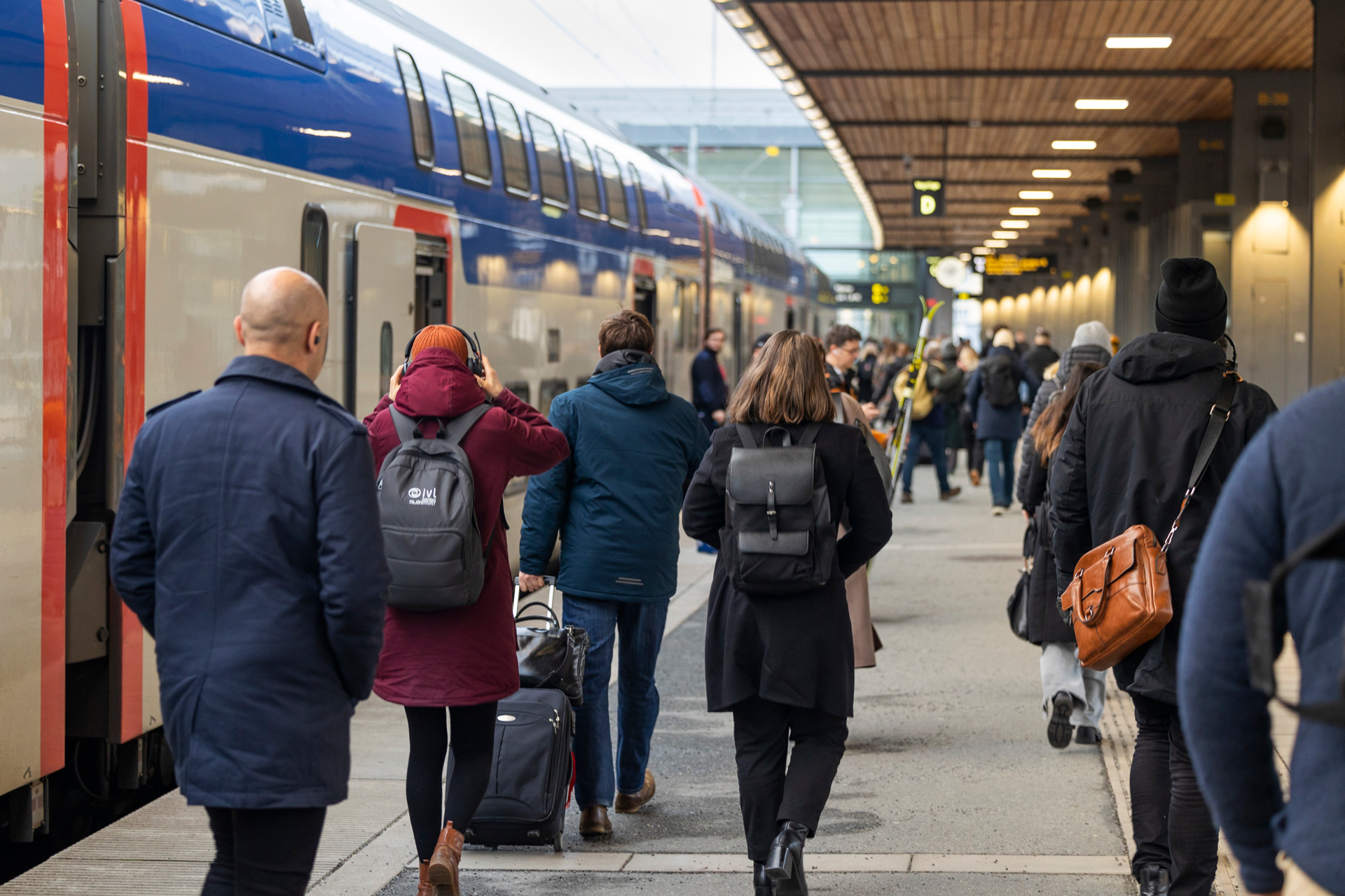Resenärer går längs en plattform bredvid ett Mälartåg på en välbelyst tågstation. Flera personer bär ryggsäckar och vinterkläder.