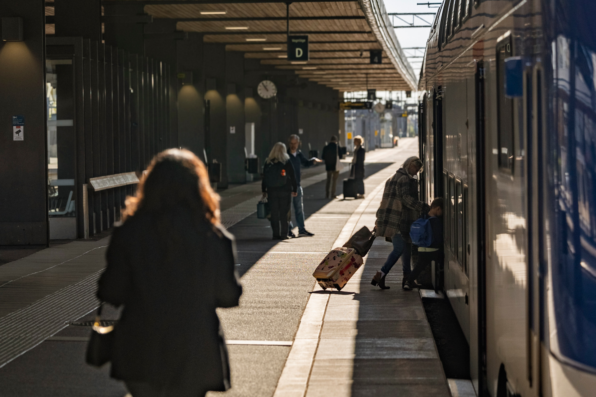  train station where several people are standing on the platform. A person with a rolling suitcase is boarding a train that is at the platform. Other people are waiting or walking along the platform.